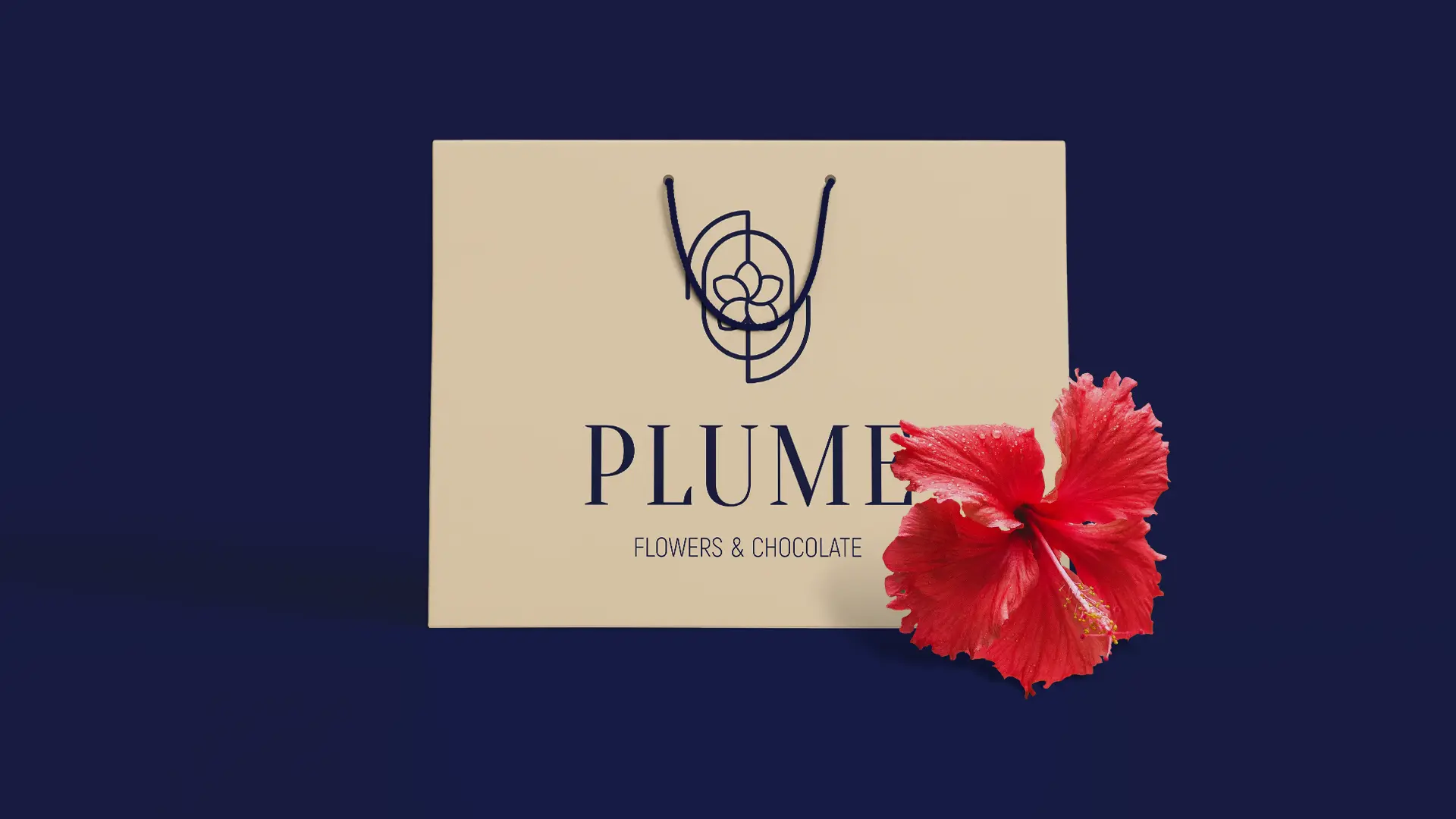 Beige Plume shopping bag with thin rope handles and the logo, set against a navy background. A decorational red hibiscus flower rests against the bag's lower right corner.